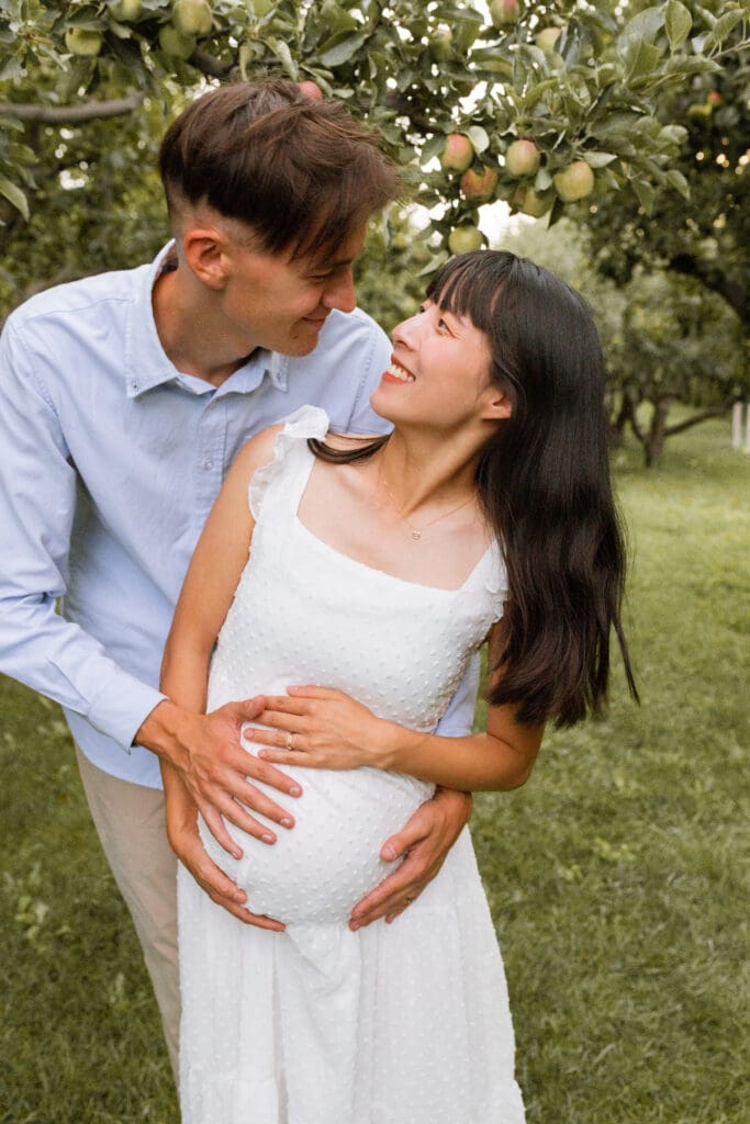 Pregnant woman cuddling with husband through apple orchard during golden hour maternity session in Texas Hill Country